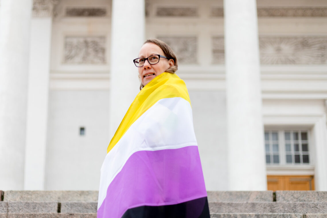 Hippu Helminen Helsingin tuomiokirkon portailla kietoutuneena kelta-valko-violettiin non-binary-lippuun.