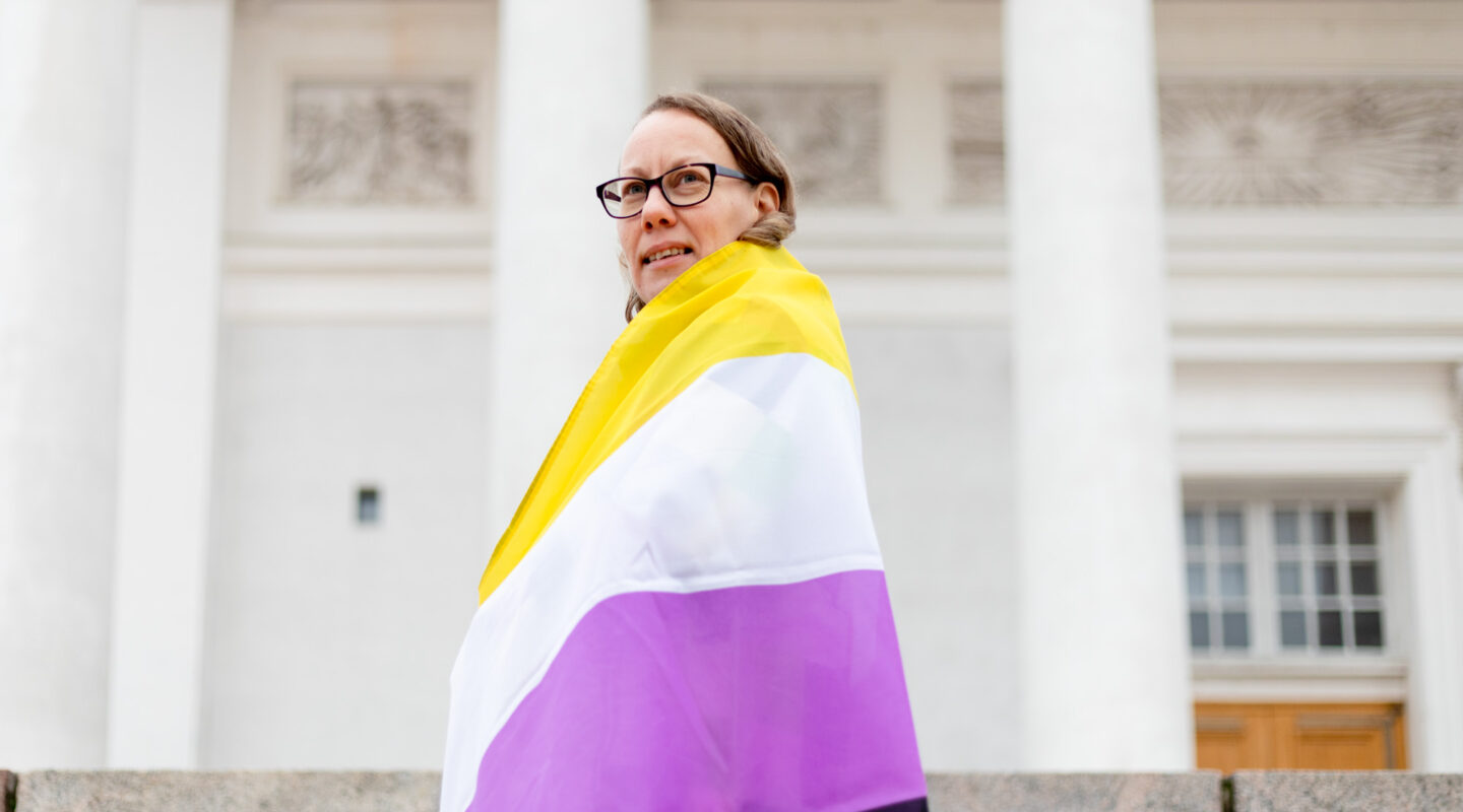 Hippu Helminen Helsingin tuomiokirkon portailla kietoutuneena kelta-valko-violettiin non-binary-lippuun.