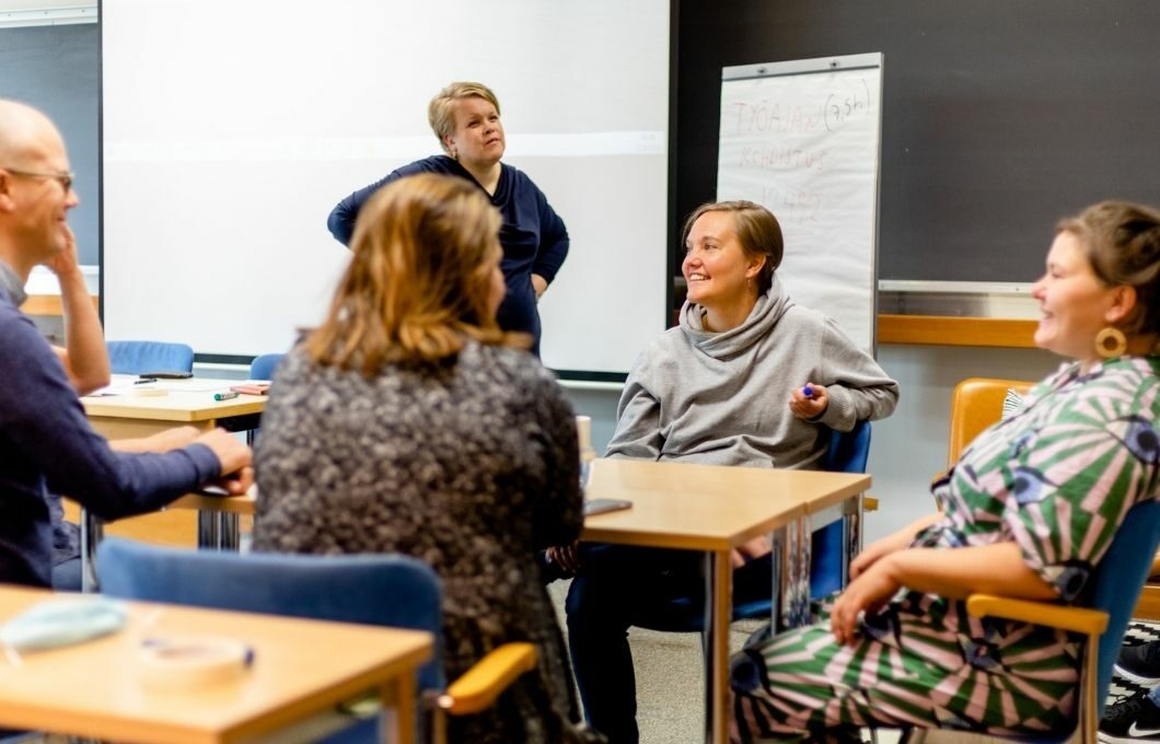 Kolme henkilöä pöydän ääressä keskustelemassa, kolmas seisoo miettivän näköisenä vieressä. Kuvan päällä on kuva Avoin osasto -oppaasta.