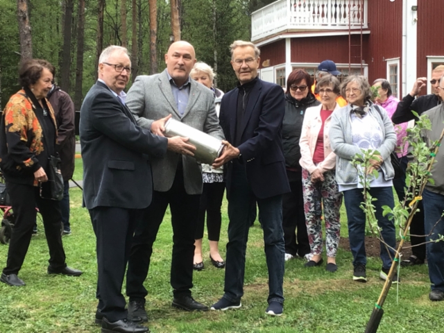 Kolme henkilöä juhlavassa asussa pitelee käsissään metallista sylinteriä puutalon pihalla. Taustalla seisoo muita tilaisuuteen osallistuvia.