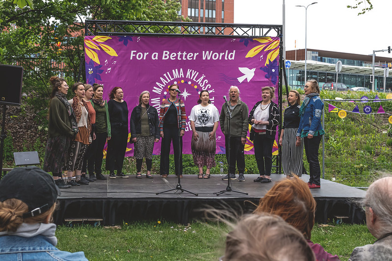 Kuoro seisoo ulkolavalla kesäisessä kaupunkimaisemassa. Lavan taustalla on lakana, jossa lukee For a better world, maailma kylässä.