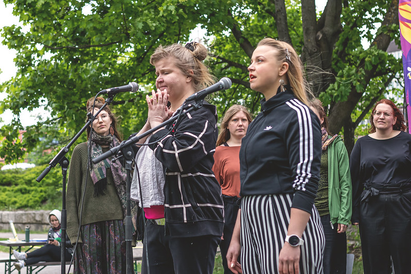 Kaksi henkilöä laulaa kesällä ulkona lavalla mikrofoniin. Taustalla näkyy lavalla muita kuorolaisia.