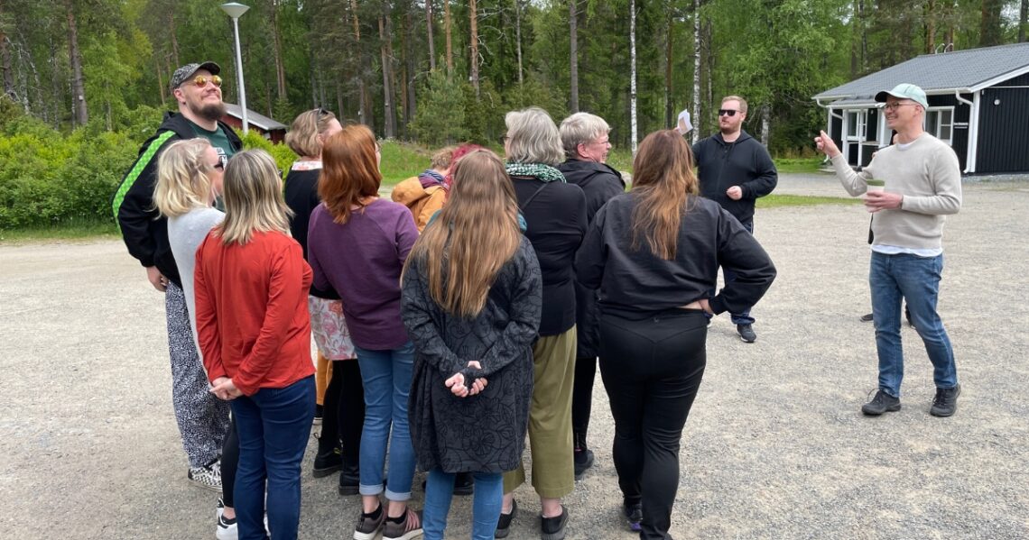 Iloinen ryhmä seisoo pihalla tiiviisti lähekkäin. Kaksi henkilöä antaa ryhmälle ohjeita.