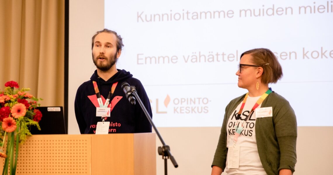 Kaksi henkilöä seisoo lavalla ja heistä toinen puhuu puhujanpöntössä. Taustalla näkyy powerpoint, jossa lukee: Kunnioitamme muiden mielipiteitä. Emme vähättele toisten kokemuksia.