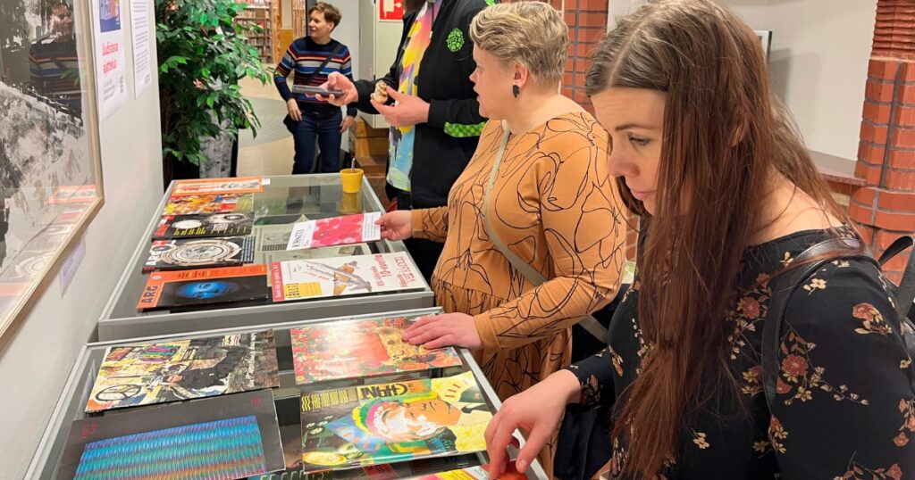 Kaksi ihmistä katselee vitriinin päällä olevia värikkäitä lehtiä. Kolmas ottaa valokuvaa vitriinistä. Neljäs katselee seinällä olevaa julistetta.