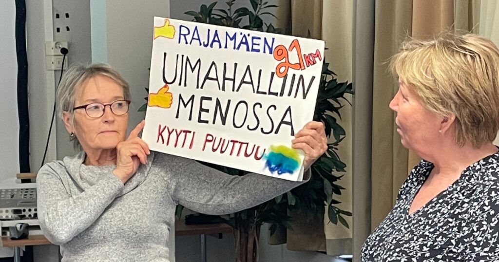 Kaksi henkilöä esittelee käsin tehtyä kylttiä, jossa lukee: Rajamäen uimahalliin menossa, kyyti puuttuu.