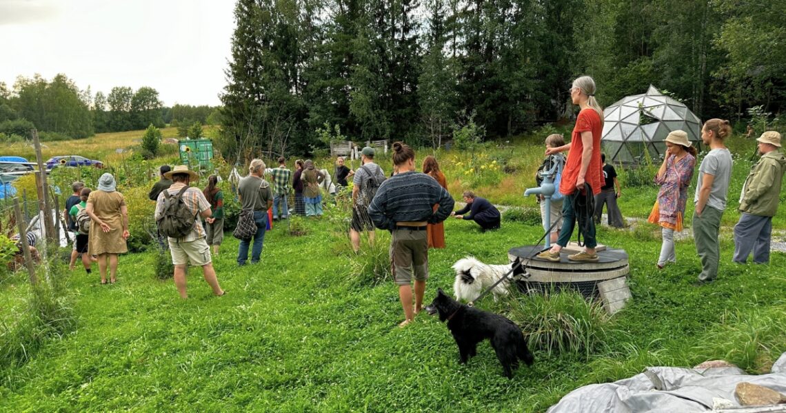 Ryhmä ihmisiä tilan pihalla katselemassa ja kuuntelemassa. Moni on pensaiden luona, yksi ottaa kyykyssä valokuvaa, kolmannella on kaksi koiraa. Ihmiset seisovat pitkäksi kasvaneella ruohikolla, taustalla näkyy metsää, kasvihuone ja muutama auto.