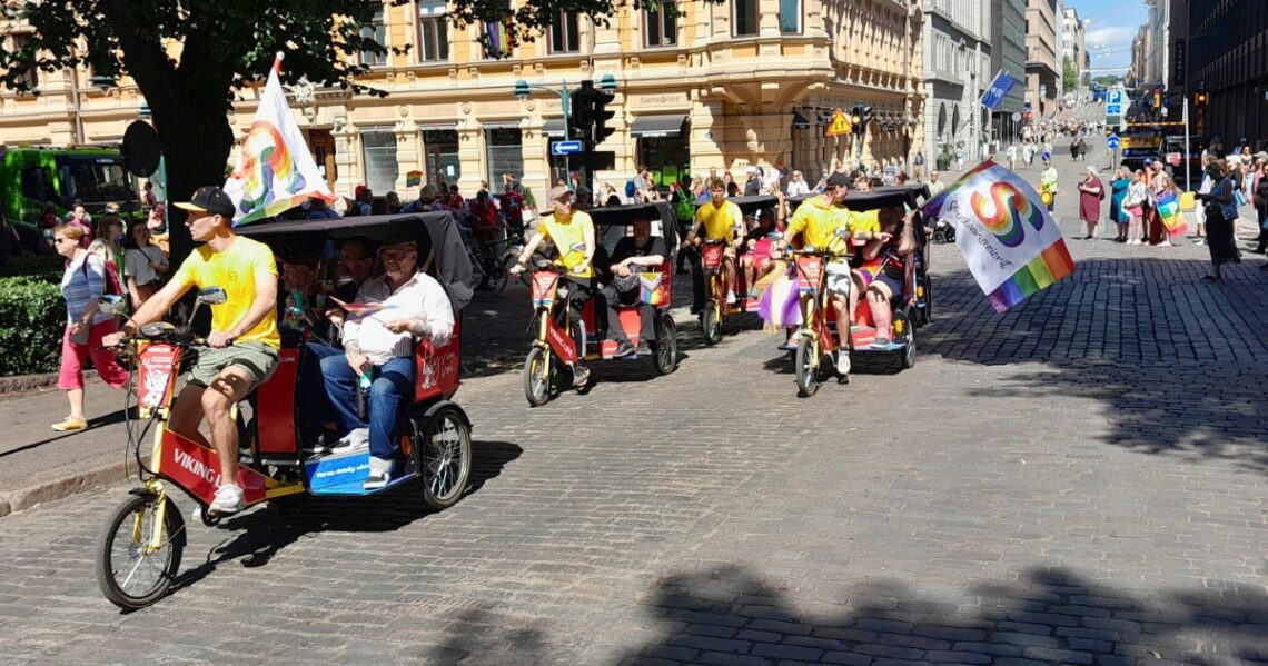 Riksha-pyöriä Helsingin Esplanadilla. Pyörien kyydissä istuu ihmisiä, jotka pitelevät kädessä Sateenkaariseniorien lippua. Taustalla on kulkue, jossa on saateenkaarilippuja.