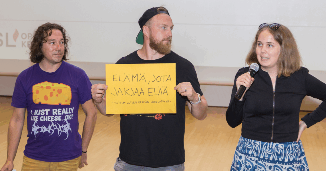 Kolme henkilöä esittelee käsinkirjoitettua kylttiä, jossa lukee: Elämä jota jaksaa elää, inhimillisen elämän edellytykset.