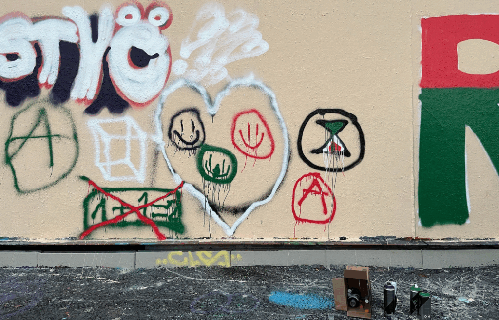 Beigelle seinälle on maalattu graffitimaaleilla erilaisia kuvioita: hymynaamoja, Elokapinan symboli, A ympyrän sisällä, laatikko.