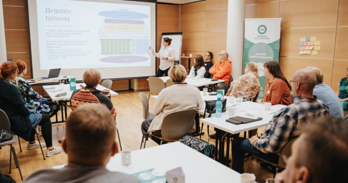 Kuva luokkatilasta, jossa kouluttaja seisoo luokan edessä ja oppijat istuvat pöydissä. Valkokankaalla on kaavio, jonka otsikko on Järjestön toiminta.
