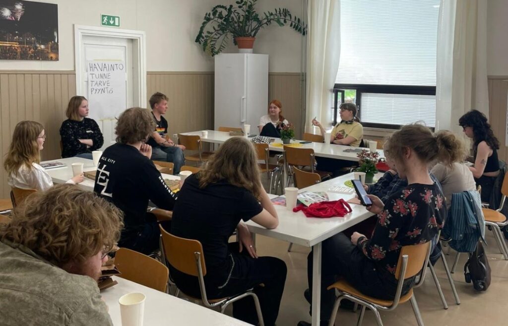 Ryhmä luokkahuoneessa keskustelemassa. Seinällä on paperi, johon on kirjoitettu Havainto, Tunne, Tarve, Pyyntö.