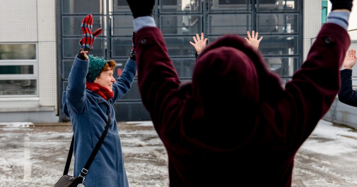 Neljä henkilöä ulkona talvisessa kaupunkitilassa parkkipaikalla. Kaikki kurottavat käsillään ilmaan.