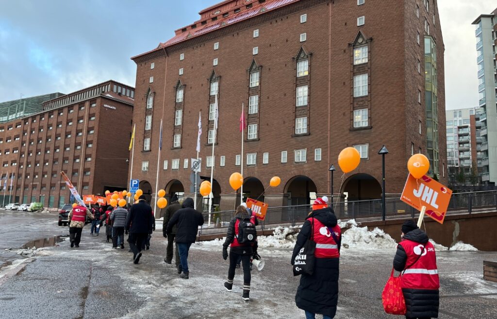 Punatiilinen kerrostalo, jonka edessä on kaariholvet. Mielenosoituskulkue kävelee rakennuksen ohi, osalla on yllään JHL:n tunnukset.