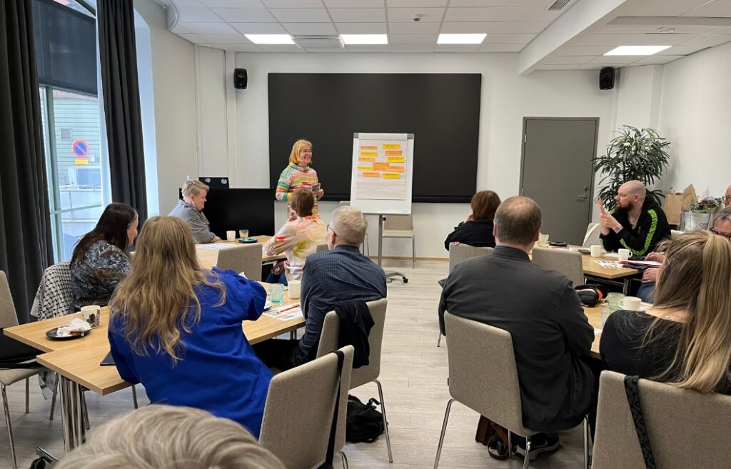 Ihmisiä istumassa pöytäkunnittain kokoustilassa, jonka etualalla seisoo kouluttaja fläppitaulun vieressä.