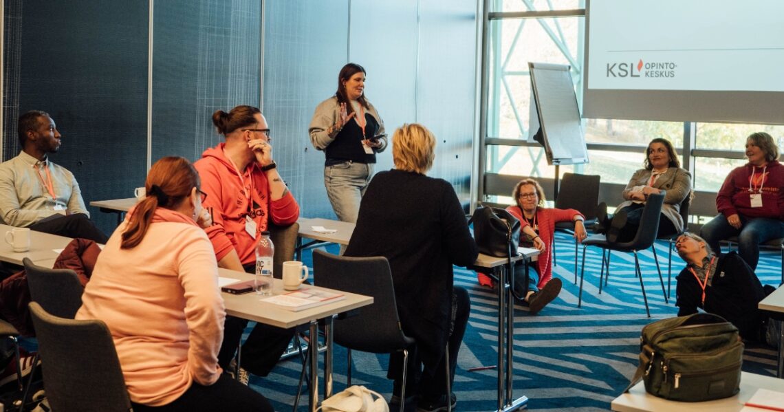 Salissa on ryhmä, jonka jäsenistä osa istuu tai makoilee rennosti lattialla kokolattiamatolla. Osa istuu pöytien ääressä katsellen edellämainittuja. Keskellä seisoo kouluttaja.