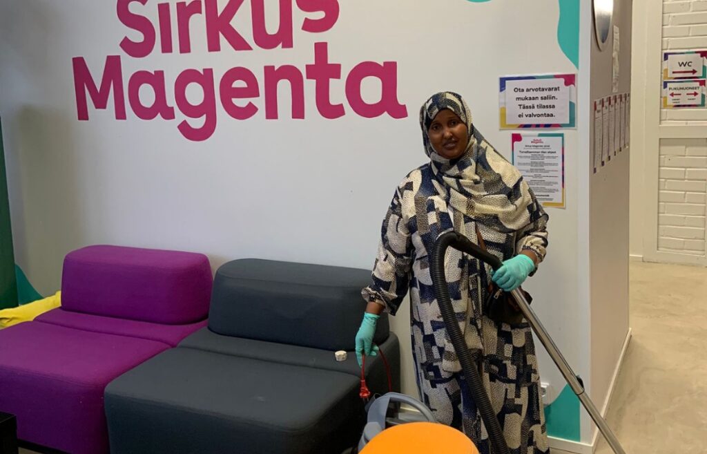 Henkilö seisoo aulassa sohvien vieressä imurin letku kädessään. Seinässä lukee teksti Sirkus Magenta.