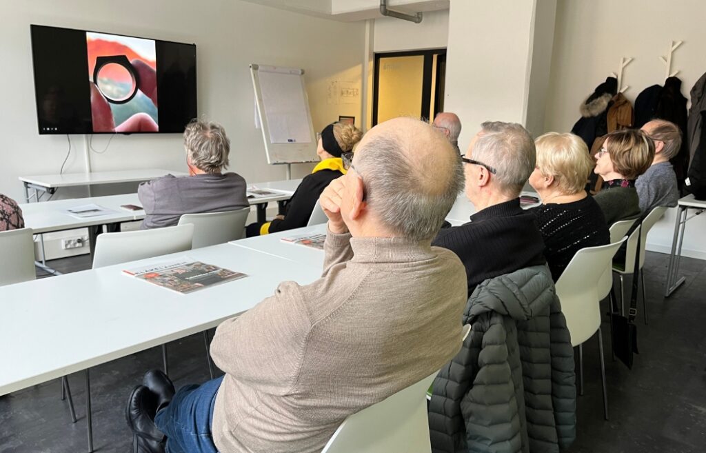 Henkilöt istuvat luokkahuoneessa ja katselevat näytöltä valokuvaa, jossa sormet pitelevät kulmikasta, sormuksennäköistä esinettä.