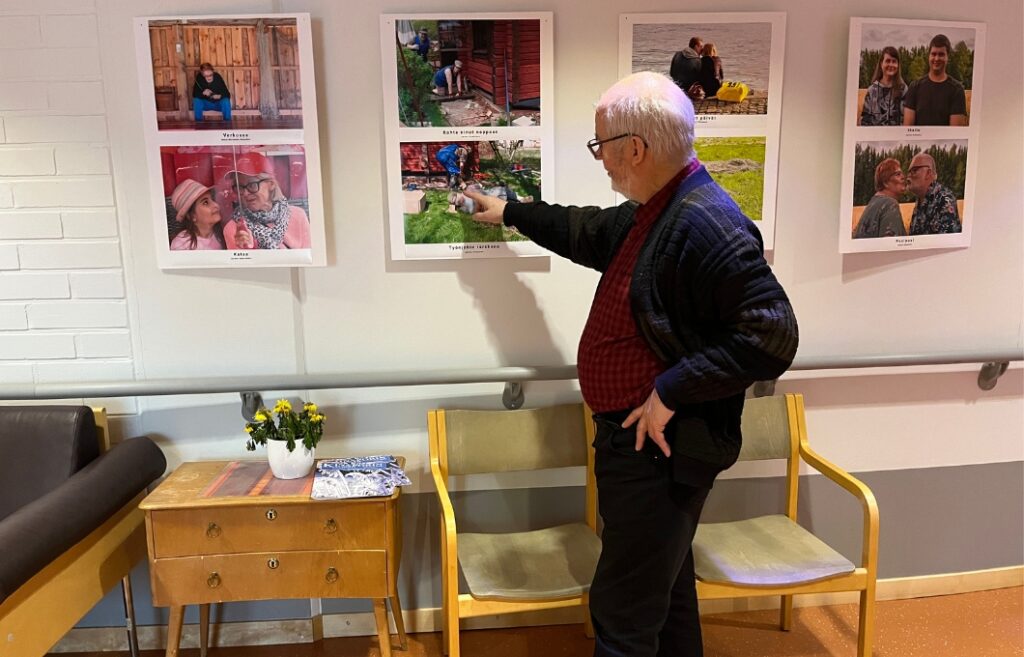 Toivo Koivisto osoittaa sormella yksityiskohtaa seinällä riippuvassa valokuvassa. Seinällä on useita valokuvia.