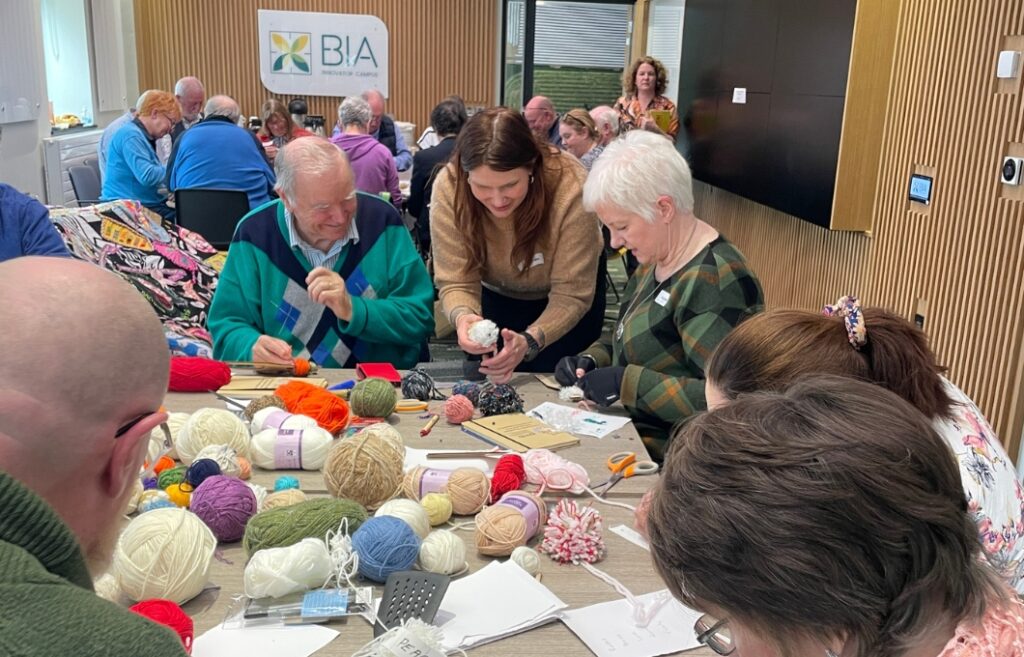 Riina Näsi kumartuneena pöydän ääreen auttamaan tupsujen tekijöitä. Pöydällä on paljon erivärisiä lankakeriä. Luokkahuoneessa istuu paljon ihmisiä.
