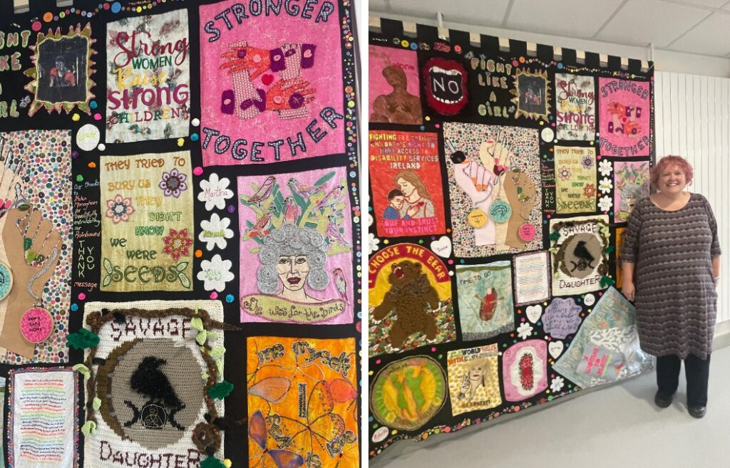 Helen Monaghan seisoo suuren seinävaatteen vieressä. Seinävaate koostuu naisten oikeuksia korostavista tilkkutöistä.