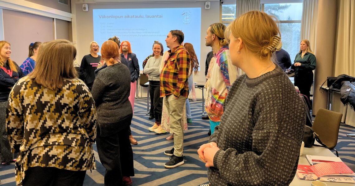 Ryhmä aikuisopiskelijoita seisoskelee luokkatilassa ja kuuntelee yhtä, joka puhuu.