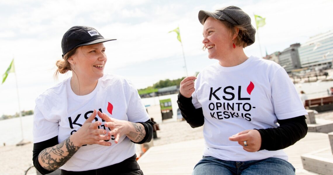 Kaksi henkilöä keskustelee iloisesti tuulisella Kauppatorilla Helsingissä.