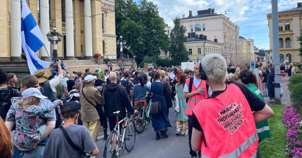 Mielenosoittajien kulkue Säätytalon edessä Helsingissä. Yksi kantaa Suomen lippua, toisella on polkupyörä. Etualalla on huomioliivissä henkilö, jonka selässä lukee häirintäyhdyshenkilö.