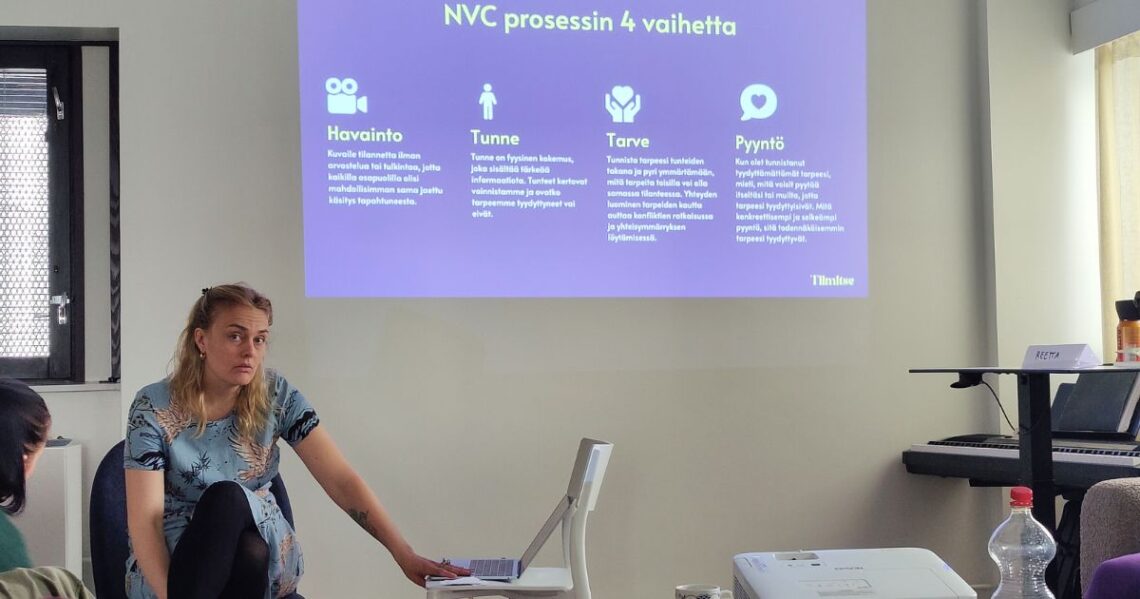 Henkilö näyttää tietokoneella luokkahuoneessa diaa, jossa lukee: NVC prosessin 4 vaihetta. Havainto. Kuvaile tilannetta ilman arvostelua tai tulkintaa, jotta kaikilla osapuolilla olisi mahdollisimman sama jaettu käsitys tapahtuneesta. Tunne. Tunne on fyysinen kokemus, joka sisältää tärkeää informaatiota. Tunteet kertovat voinnistamme ja ovatko tarpeemme tyydyttyneet vai eivät. Tarve. Tunnista tarpeesi tunteiden takana ja pyri ymmärtämään, mitä tarpeita toisilla voi olla samassa tilanteessa. Yhteyden luominen tarpeiden kautta auttaa konfliktien ratkaisussa ja yhteisymmärryksen löytämisessä. Pyyntö. Kun olet tunnistanut tyydyttömättömät tarpeesi, mieti, mitä voisit pyytää itseltäsi tai muilta, jotta tarpeesi ttyydyttyisivät. Mitä konkreettisempi ja selkeämpi pyyntö, sitä todennäköisemmin tarpeesi tyydyttyvät. Tiimitse.