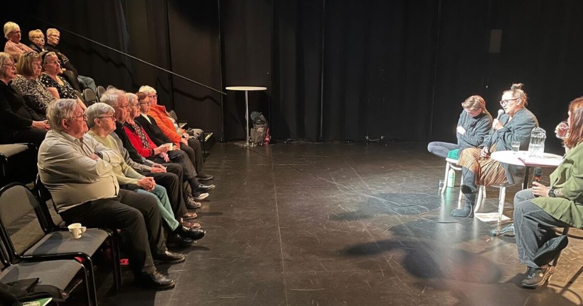 Valokuva teatterista. Yleisöä istuu teatterin katsomossa keskustelemassa lavalla istuvien kolmen henkilön kanssa.